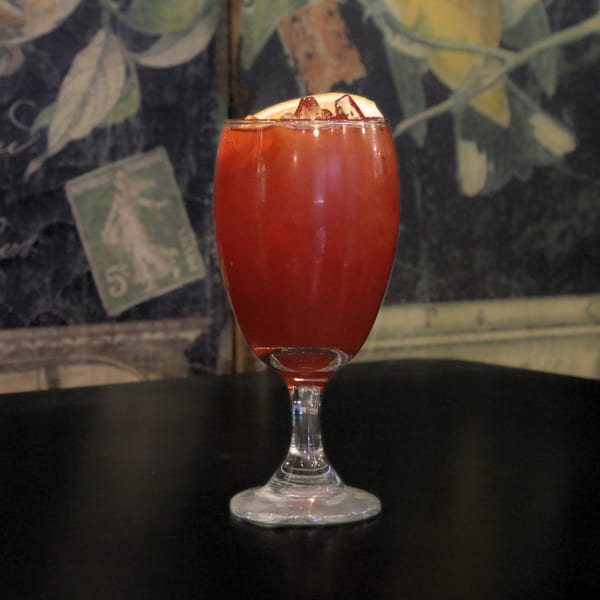 A vibrant red cocktail with ice and an orange slice, on a dark table against a patterned wall.
