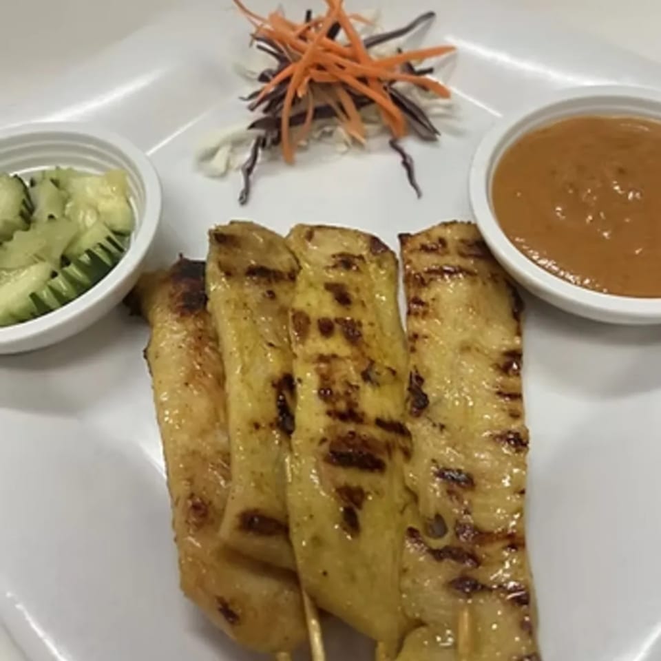 Grilled chicken satay skewers with peanut sauce, cucumber salad, and shredded carrots on a white plate.