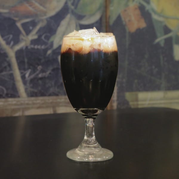 Iced Thai coffee with a creamy top layer and ice in a stemmed glass on a dark table.