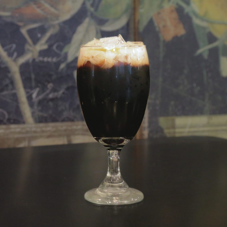 Iced Thai coffee with a creamy top layer and ice in a stemmed glass on a dark table.