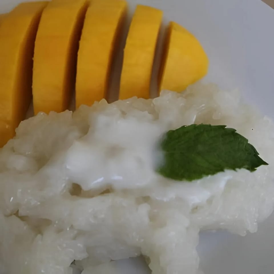 Mango sticky rice with coconut milk and a mint leaf on a white plate.