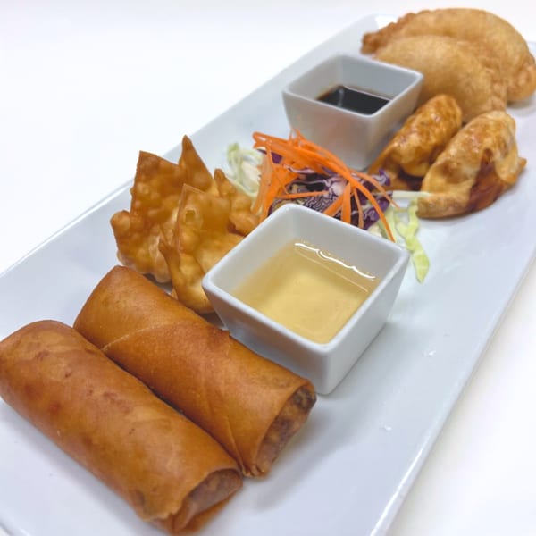 A white rectangular plate holds spring rolls, fried wontons, and dumplings with two dipping sauces.