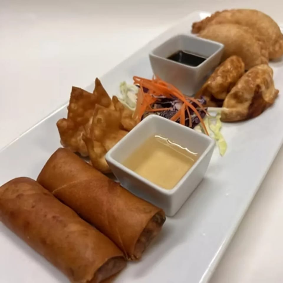 A white rectangular plate holds spring rolls, fried wontons, and dumplings with two dipping sauces.